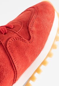 Punaiset mokkanahkakengät, joissa on rakennepintainen yläosa, kontrastoiva valkoinen välikenkä ja kuminen ulkopohja, jossa on näkyvät pito-urat.