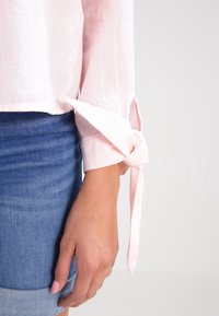 Chemisier en tissu rose clair côtelé avec un détail de manche nouée, associé à une jupe en denim bleu. Gros plan sur la manche et la main.