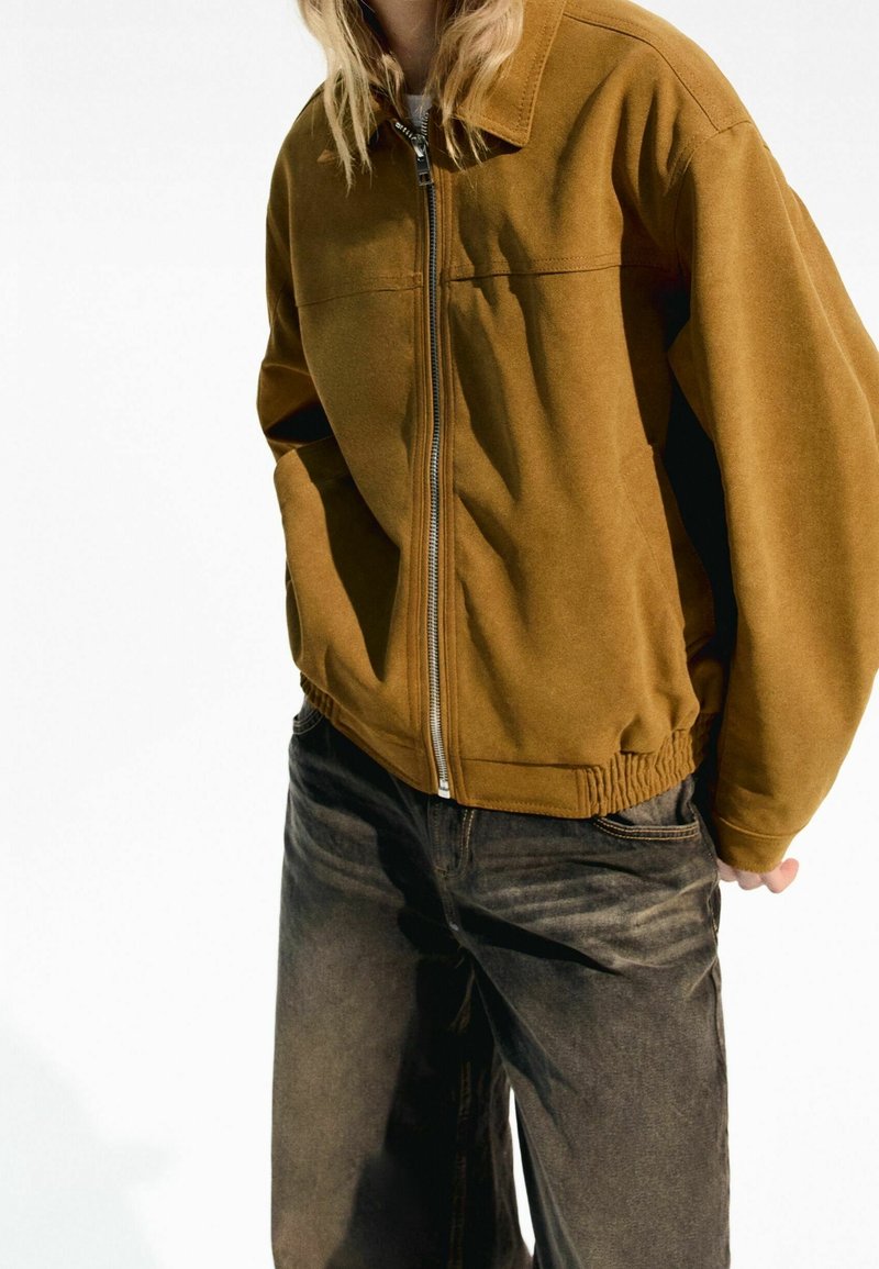 Veste marron à fermeture éclair avec col chemise, taille élastiquée et texture lisse, accompagnée d'un pantalon en denim foncé.