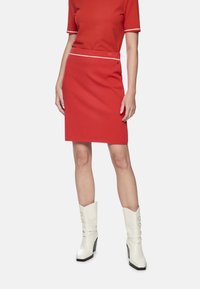 Rotes, figurbetontes Kleid mit kurzen Ärmeln, weißem Besatz und passendem Taillenband. Kombiniert mit weißen Knöchelstiefeln mit klobigen Absätzen und goldenen Akzenten.