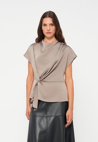 Top beige drappeggiato con maniche corte, caratterizzato da un dettaglio a nodo in vita; texture liscia e caduta fluida. Indossato con una gonna di pelle nera.
