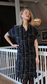 Sort skjortekjole med hvide geometriske mønstre, korte ærmer, knapper foran og krave. Stoffet har en glat struktur og falder til midt på læggen.