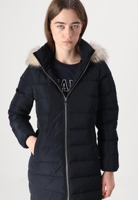 Cappotto puffer blu navy con cappuccio con bordo in pelliccia, chiusura con zip e tasche laterali. Presenta un design trapuntato e una texture morbida.