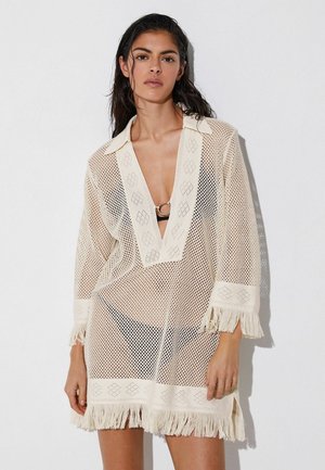 Femeie purtând o vestă crem din plasă cu margine cu franjuri peste un costum de baie negru, stând în fața unui perete alb simplu.