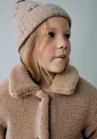 Chaqueta beige de felpa con un gran cuello y cierre de botones, combinada con un gorro de punto ribeteado beige que tiene un pompón en la parte superior.