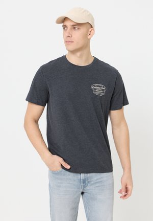 Mann mit beigefarbener Mütze, dunkelgrauem T-Shirt mit Grafik und hellblauen Jeans, der nach links schaut und eine Hand in der Tasche hat.