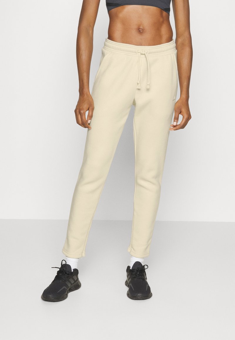 Pantalones de chándal color crema con un ajuste tapered, cintura con cordón y dobladillos ajustados, combinados con zapatillas deportivas negras y calcetines blancos de caña baja.