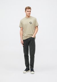 Beige puuvillane T-särk, millel on must logo prinditud, koos tumedate hallide teksade ja mustade tossudega valgete talladega, lihtsal taustal.