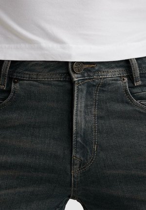 Krupni plan tamnoplave teksas traperice s gumbom zakopčanim, vidljivim petljama za remen i običnom bijelom košuljom uvučenom unutra.