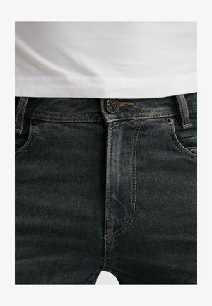 Krupni plan tamnoplave teksas traperice s gumbom zakopčanim, vidljivim petljama za remen i običnom bijelom košuljom uvučenom unutra.