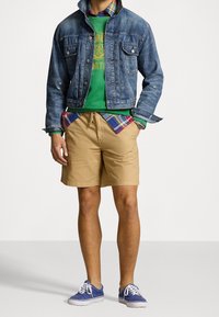 Hombre vestido con una chaqueta de mezclilla azul, sudadera verde, camisa a cuadros debajo, pantalones cortos caqui y zapatillas de lona azules, de pie con las manos en los bolsillos.