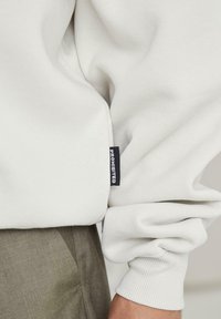 Felpa bianca con polsini a costine, caratterizzata da un piccolo etichetta nera con la scritta "PROIBITO". Indossata sopra pantaloni di tessuto verde, che mettono in mostra un look casual.