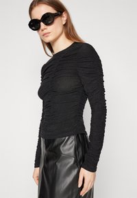Svart langermet topp med samlet tekstur; parret med et svart skinnskjørt. Modellen har store, runde svarte solbriller.