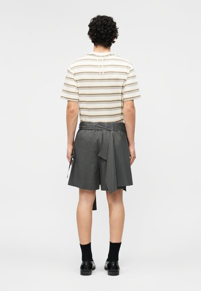 Uomo con maglietta a righe crema e marrone a maniche corte, pantaloncini grigi larghi a vita alta, calzini neri e scarpe eleganti nere, in piedi di spalle.