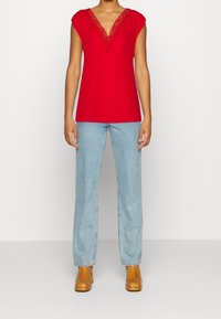 Punainen paita syvällä V-kaula-aukolla, jossa pitsireunus, yhdistettynä vaaleansinisiin farkkuihin ja ruskeisiin nilkkabootseihin, eteenpäin katsottuna yksinkertaista taustaa vasten.