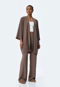 Ensemble en maille brun texturé comprenant un long cardigan ouvert et un pantalon large, associé à un top ajusté blanc et des sandales.