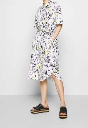 Woman wearing a white knee-length shirt dress with black and yellow floral patterns, paired with black platform slide sandals.