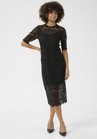 Vestido de encaje negro con mangas cortas, que presenta patrones intrincados, una silueta ajustada y un dobladillo con forma de ondulación. Combinado con zapatos de tacón negros.