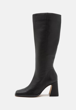 Sort læderstøvle til midt på læggen med firkantet tå og kraftig blokhæl på hvid baggrund, sidevisning.