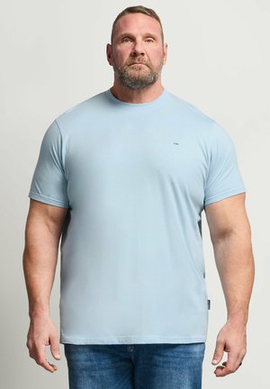 Skægget mand med kort hår står vendt fremad iført en lyseblå T-shirt og blå jeans mod en ensfarvet lys baggrund.