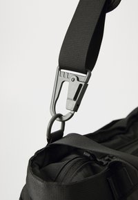 Bolsa de tela negra con correa ajustable y mosquetón de plástico gris. Material texturizado con forma delgada y múltiples compartimentos con cremallera.