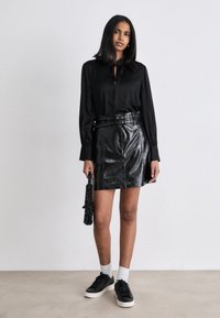 Blusa nera con zip frontale e laccio, abbinata a una mini gonna in pelle nera con vita alta e tasche. Scarpe da ginnastica nere e borsa strutturata.