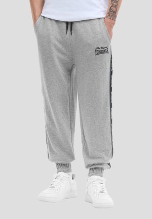 Persoon draagt lichtgrijze Lonsdale joggingbroek met zwarte strepen aan de zijkant, gecombineerd met witte sneakers, handen in de zakken, zichtbare getatoeëerde arm.