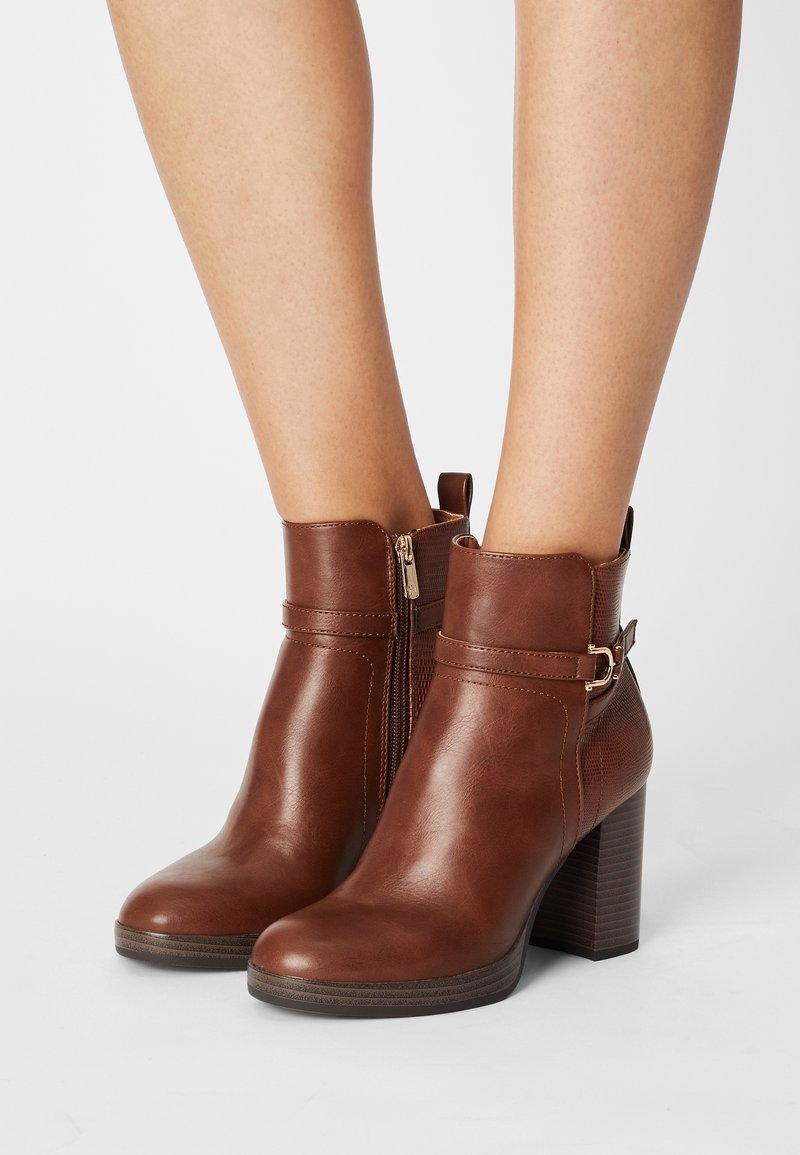 Bottes cuir marron à la cheville avec fermeture éclair latérale, boucle décorative et talons blocs portées sur jambes nues sur un fond uni.