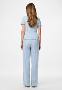 Ženska z dolgimi valovitimi lasmi, oblečena v svetlo modro kratkodlačno majico in ujemajoče široke hlače, stoji obrnjena stran od bele ozadja.