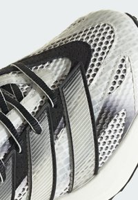 Sportig sko med ett texturerat svartvitt meshtyg på ovansidan, med tre silverfärgade ränder och svarta snören. Gummisula med strukturerad design.