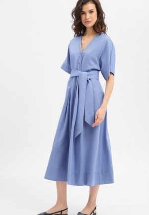 Vrouw die een lichtblauwe midi-jurk draagt met brede korte mouwen en een brede, gestrikte ceintuur, staand tegen een effen witte achtergrond.