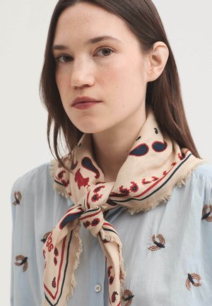 Junge Frau mit braunen Haaren trägt einen beigen Schal mit roten und blauen Paisley-Mustern über einer hellblauen bestickten Bluse.