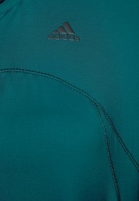 Tyrkysové sportovní tričko s hladkým, elastickým materiálem. Obsahuje prošívané detaily a malé černé logo Adidas na levém horním rohu.