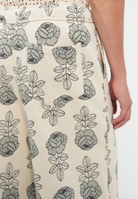 Pantalones de algodón beige con un patrón floral en gris, con un bolsillo parche en la parte posterior y detalles de textura sutiles.