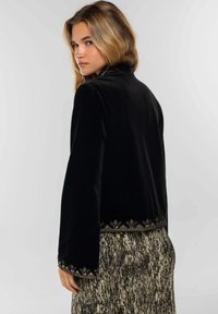 Veste en velours noir à manches longues, ornée d'une broderie florale métallique dorée sur l'ourlet. Portée sur une jupe texturée, mettant en valeur un look à couches.