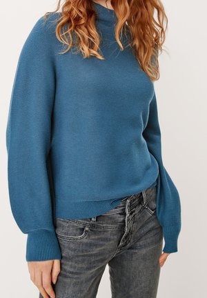 Vrouw draagt een blauwe groenen gebreide trui met lange mouwen met omgeslagen manchetten en verweerde zwarte high-waisted jeans.