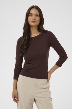 Mujer con cabello largo y oscuro, vistiendo una blusa marrón de manga tres cuartos y pantalones beige, posando con una mano en la cadera.