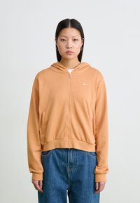 Oransje hettejakke med glidelås, laget av myk stoff. Har sidelommer og en liten hvit logo på brystet. Bært med blå jeans.