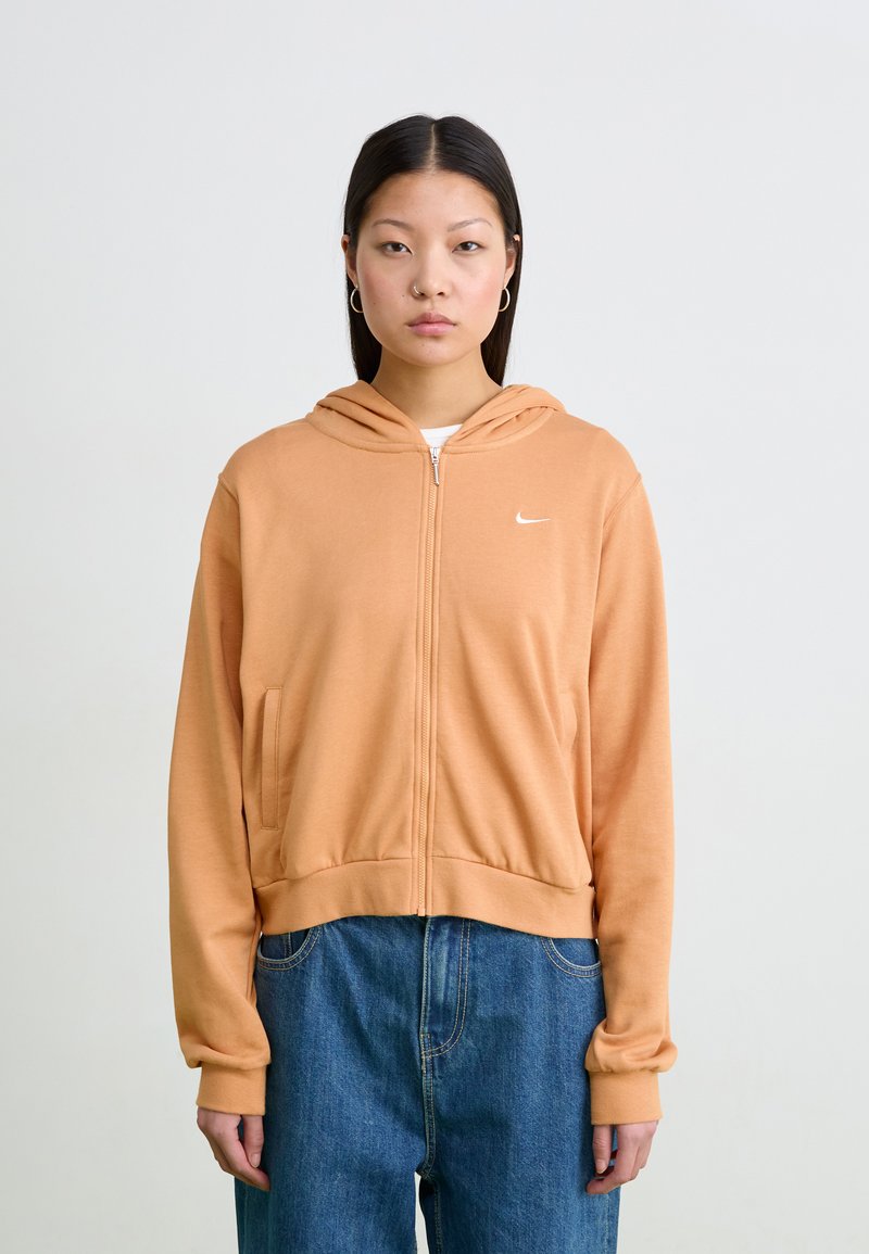 Oransje hettejakke med glidelås, laget av myk stoff. Har sidelommer og en liten hvit logo på brystet. Bært med blå jeans.