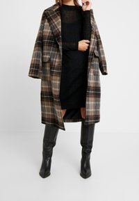 Rutig ullkappa i brunt och grönt, svart nätkjol med lång ärm under, kombinerat med figursydda svarta läderstövlar som går upp till knäna.