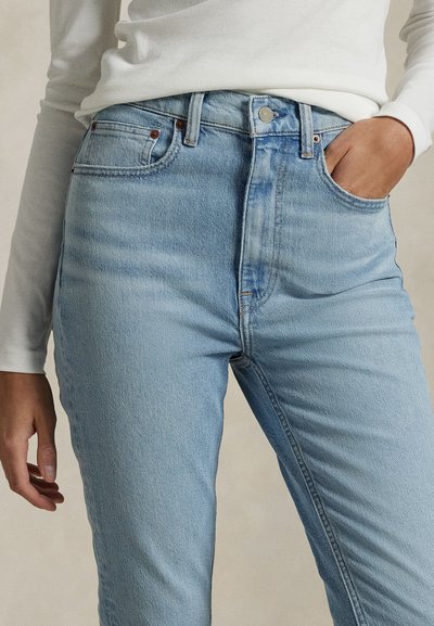 Persoon draagt lichtblauwe high-waisted jeans en een wit top met lange mouwen, met één hand in een zak en de andere ontspannen langs het lichaam.