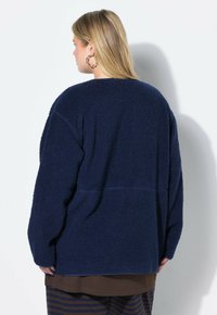 Blonde Frau, die große Kreolenohrringe und eine strukturiert navyblaue Fleecejacke trägt, von hinten vor einem weißen Hintergrund gezeigt.