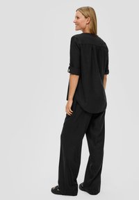 Chemise en lin noir, ample, avec des manches retroussées et un ourlet arrondi, associée à un pantalon large noir et des sandales noires. Design simple, sans motifs.