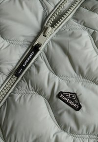 Lichtgroene gewatteerde jassenstof met een opvallende rits, voorzien van een zwart Superdry logo patch en gestikt detail.