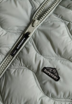 Veste matelassée vert clair avec une fermeture éclair bien en évidence, arborant un patch logo Superdry noir et des détails brodés.