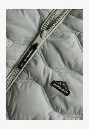 Veste matelassée vert clair avec une fermeture éclair bien en évidence, arborant un patch logo Superdry noir et des détails brodés.