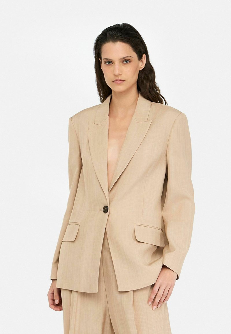 Blazer beige con cierre de un solo botón, solapas muescadas y dos bolsillos frontales. Fabricado en una tela texturizada con un corte ajustado.
