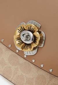 Vaaleanruskea nahkakäsilaukku, jossa on teksturoidut alaosat; varustettu kultaisella kukkaketjulla, jossa on hopeisia yksityiskohtia ja koristeellisia niittejä.