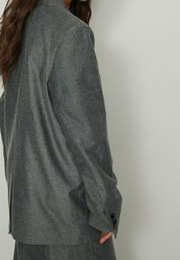 Harmaa tweed-takki, jossa on muotoiltu istuvuus, ryhdikkäät hartiat, kevyt kiilto ja pitkät hihat. Varustettu nappi-hihansuilla ja sivuhalkioilla.