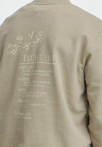 Sweatshirt bej cu un grafic imprimat "Lista de dorințe" care include destinații de călătorie și activități în alb, cu pictograme stil doodle.